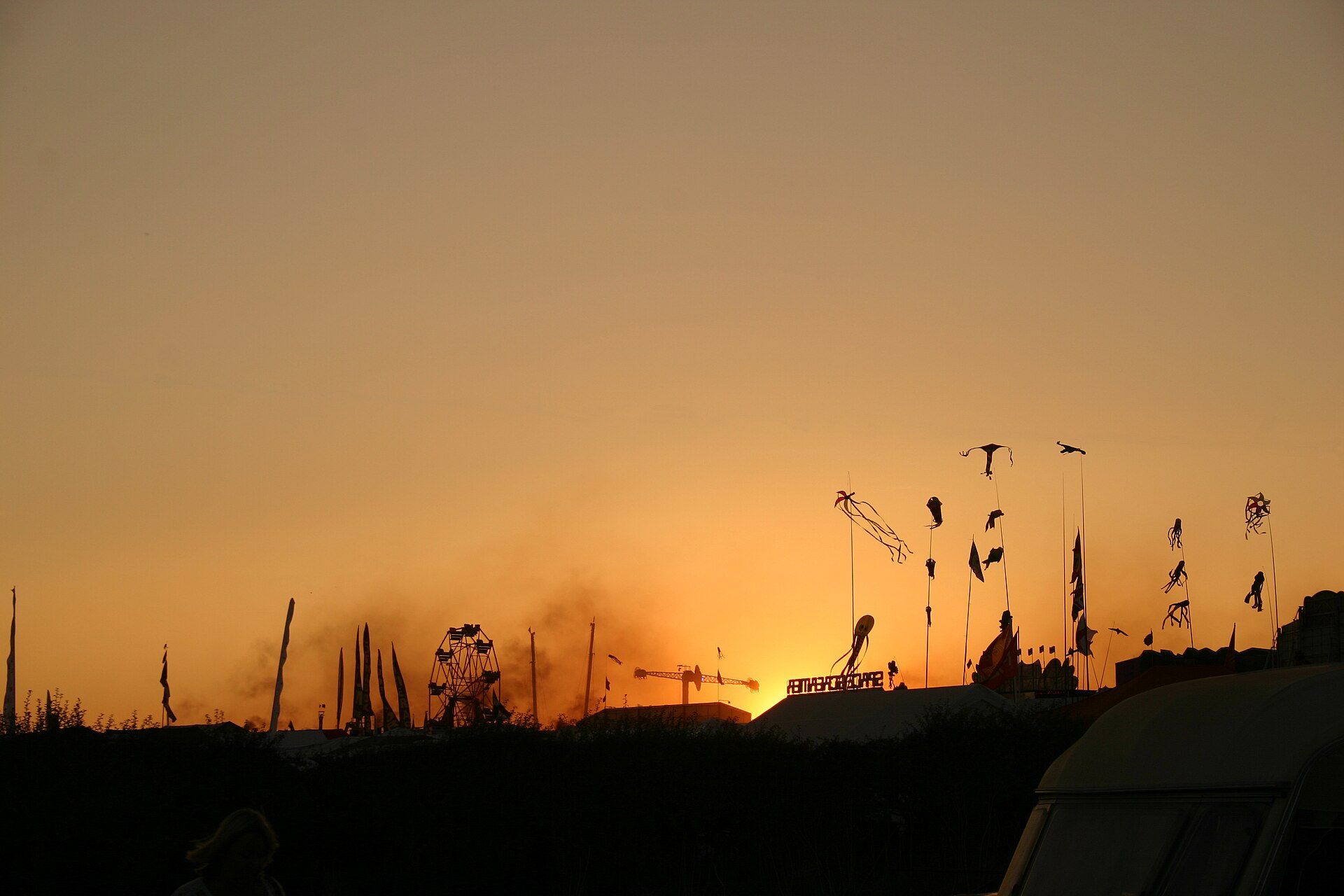 Orange sunset over the Great Dorset Steam Fair, with a big wheel, flags and kite streamers silhouetted along the skyline and haze drifting across the showground.