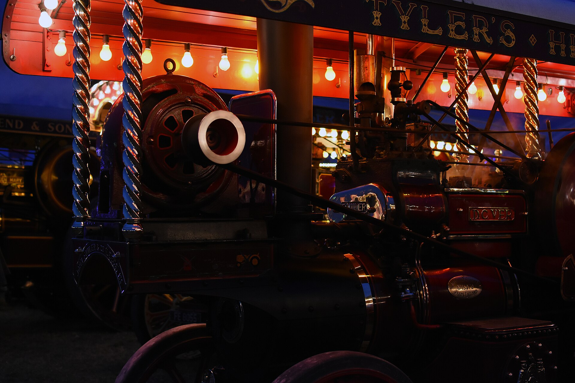 Close view of a dark red showman's steam engine at night, its brass twisted canopy columns lit by rows of warm festoon bulbs, with a deep blue dusk sky behind.