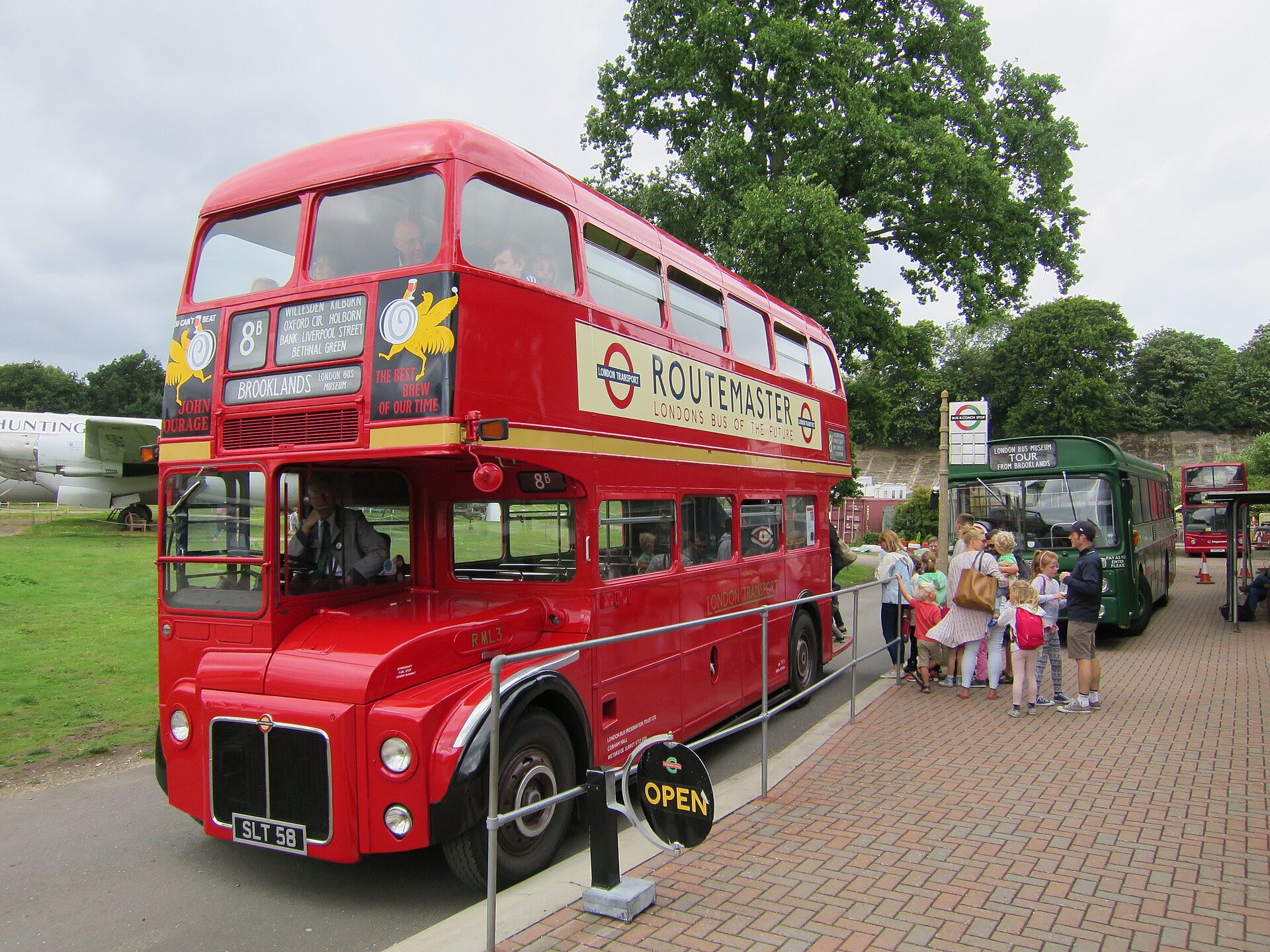 Red prototype Routemaster bus at Brooklands museum, driver in cab, children and parents queueing to board a green single-decker tour bus behind it