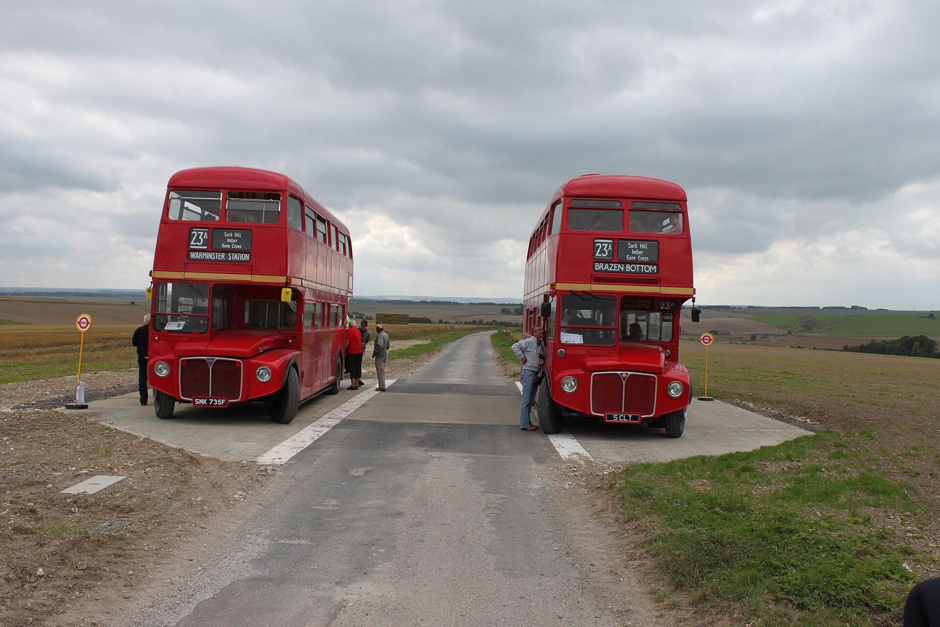 Two red Routemaster double-deckers facing each other on a concrete crossing point in open downland, with passengers standing beside them under a dramatic cloudy sky