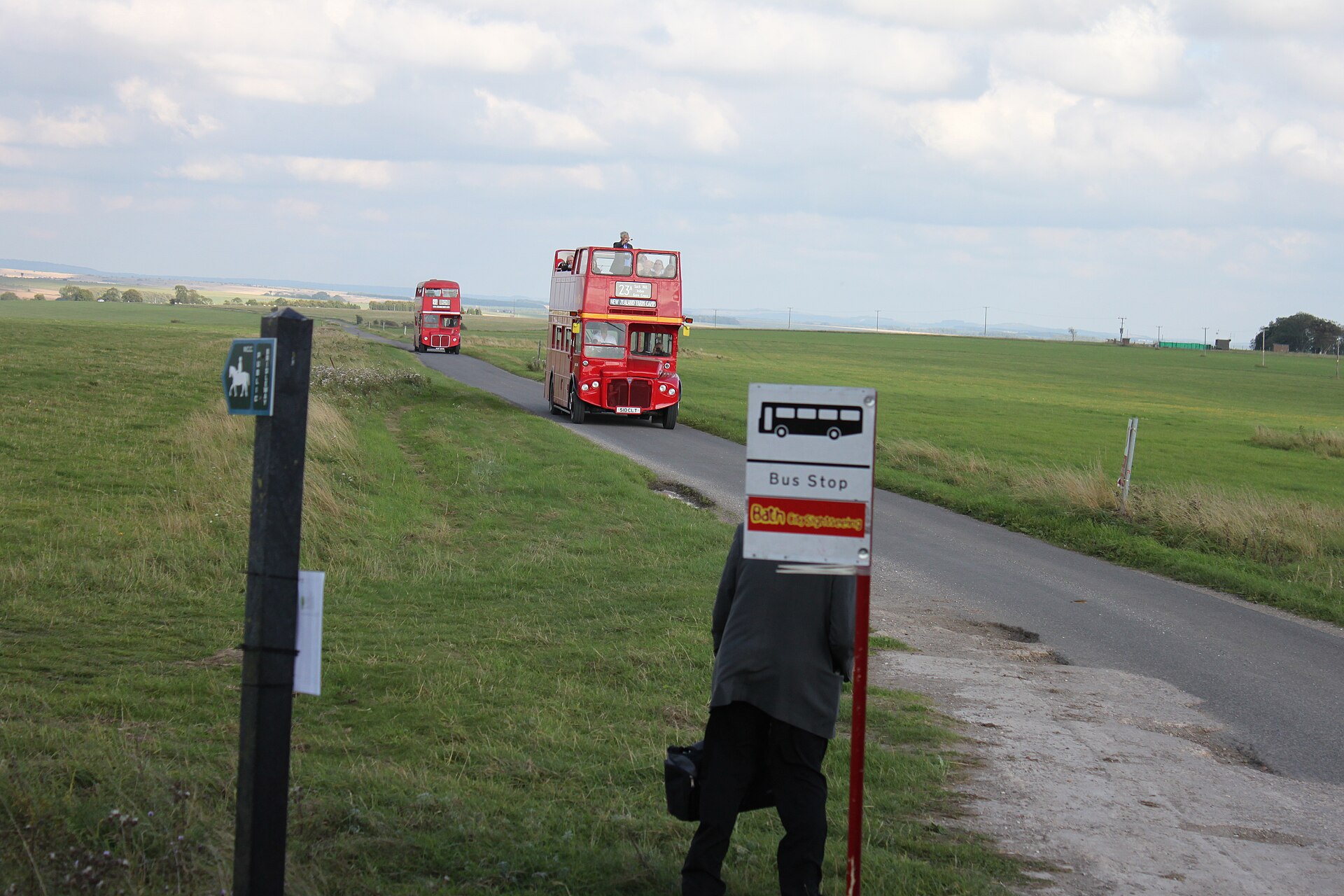 Two red Routemaster buses, the leading one open-top with passengers upstairs, coming along a narrow rural road towards a bus stop where a person waits
