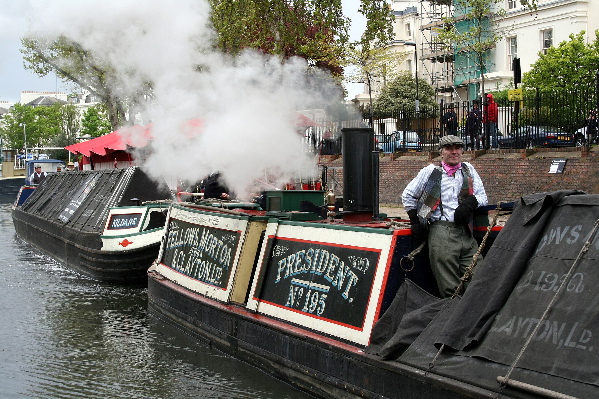 The historic steam narrowboat President billowing white steam at a canal festival, with a boatman in period waistcoat and flat cap leaning on the cabin and the butty Kildare moored alongside.