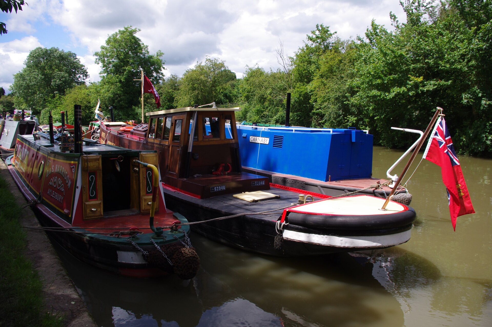 Historic canal tugs and working boats moored two abreast waiting for a rally parade, a red ensign flying from a stern mast under a bright cloudy sky.