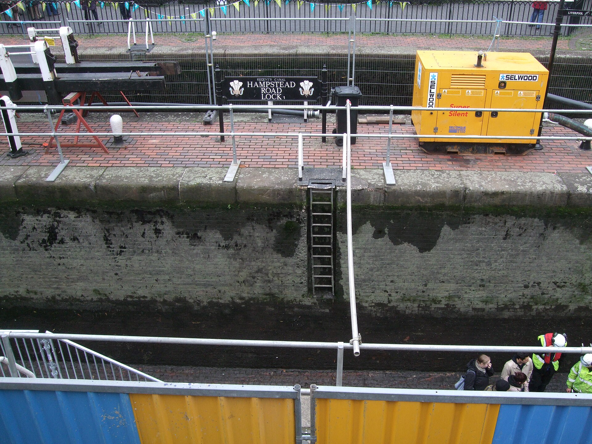 A drained canal lock chamber at Camden with the Hampstead Road Lock sign, an access ladder down the exposed brick wall, bunting overhead and a pump generator on the lockside.