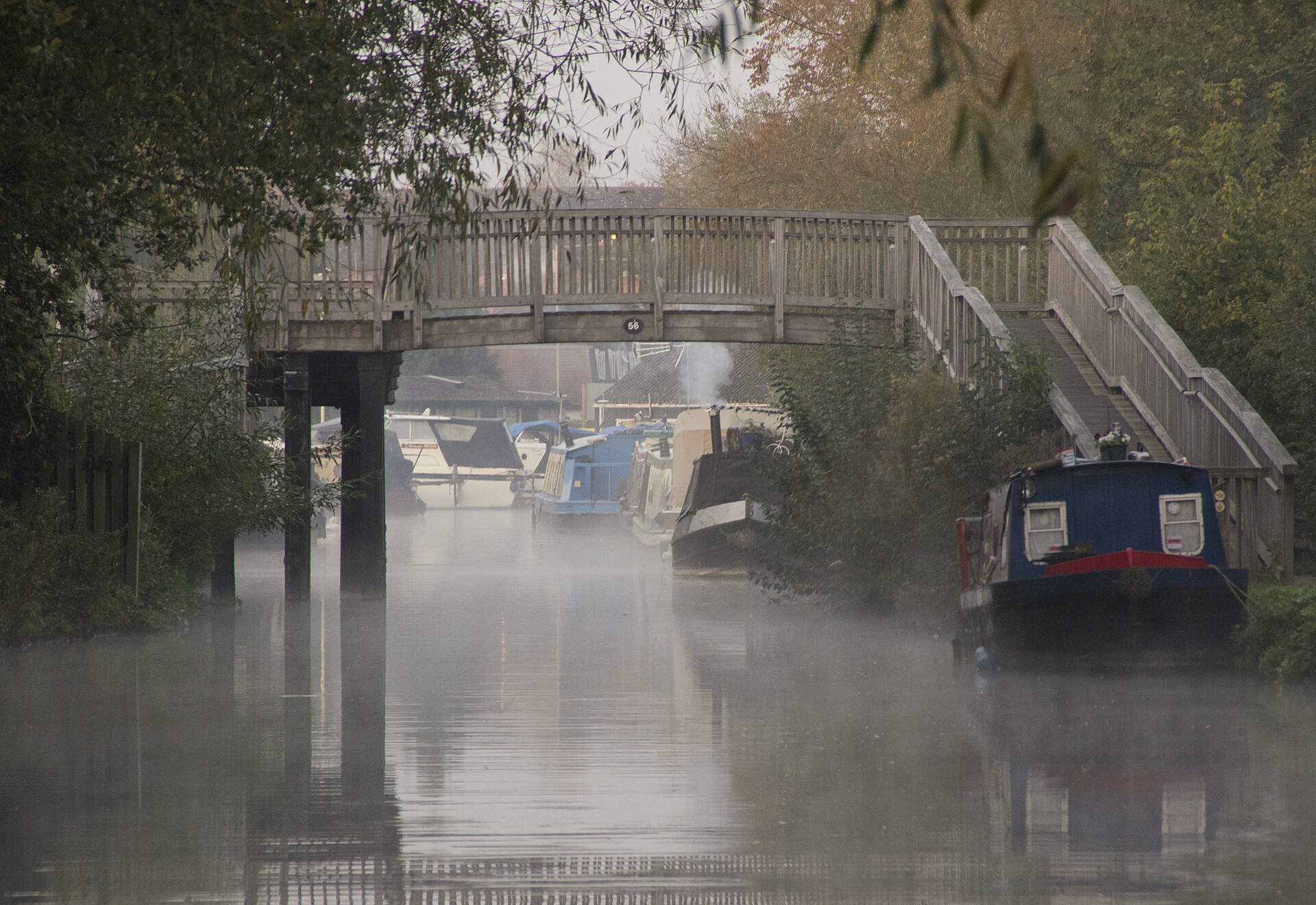 Mist hanging over a still canal at dawn beneath a wooden footbridge, moored narrowboats fading into the haze with woodsmoke rising from a stove chimney.