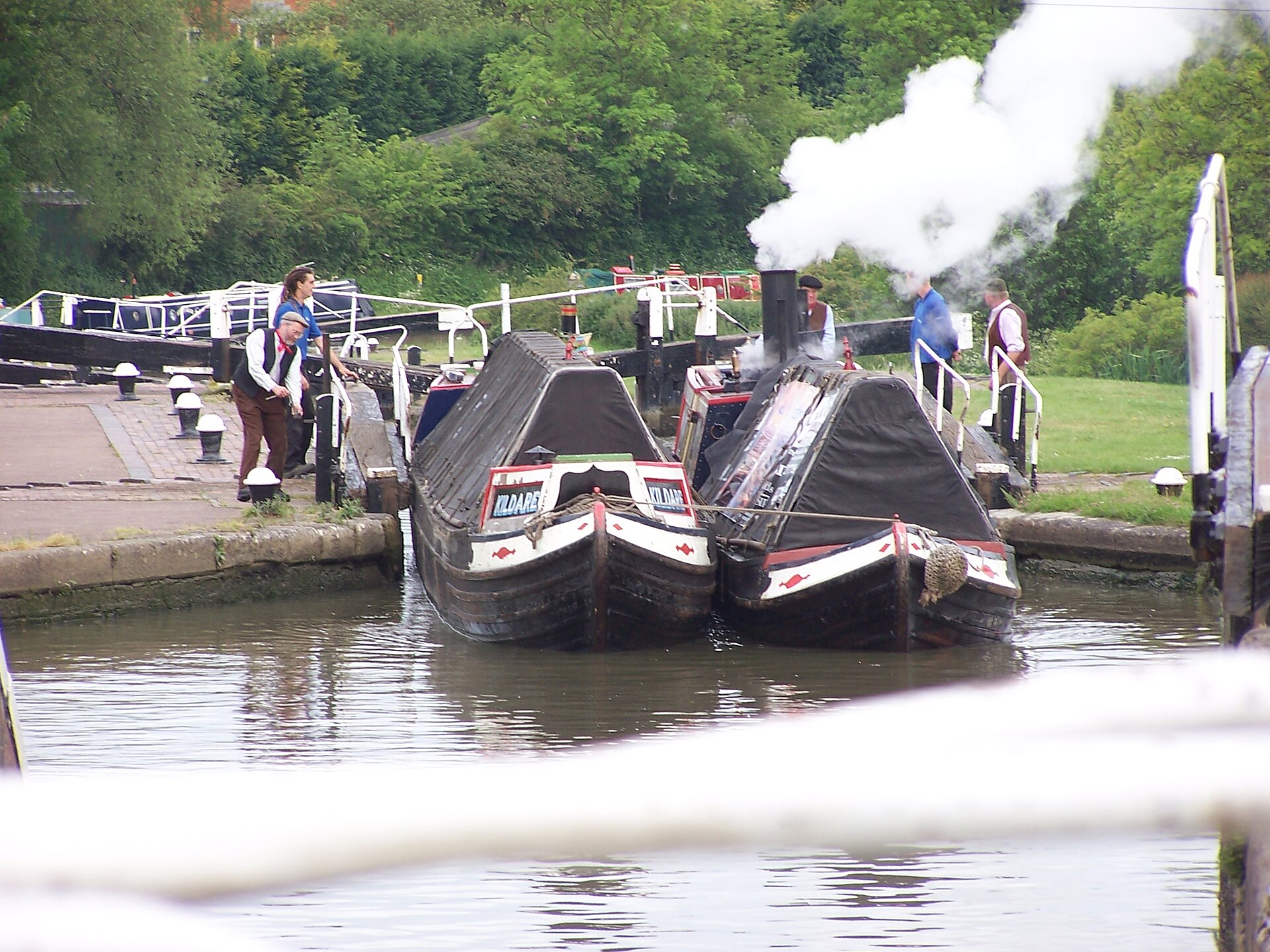 Two historic working narrowboats breasted up and leaving a canal lock together, steam rising from the motor boat's chimney while crew in traditional dress work the lockside.