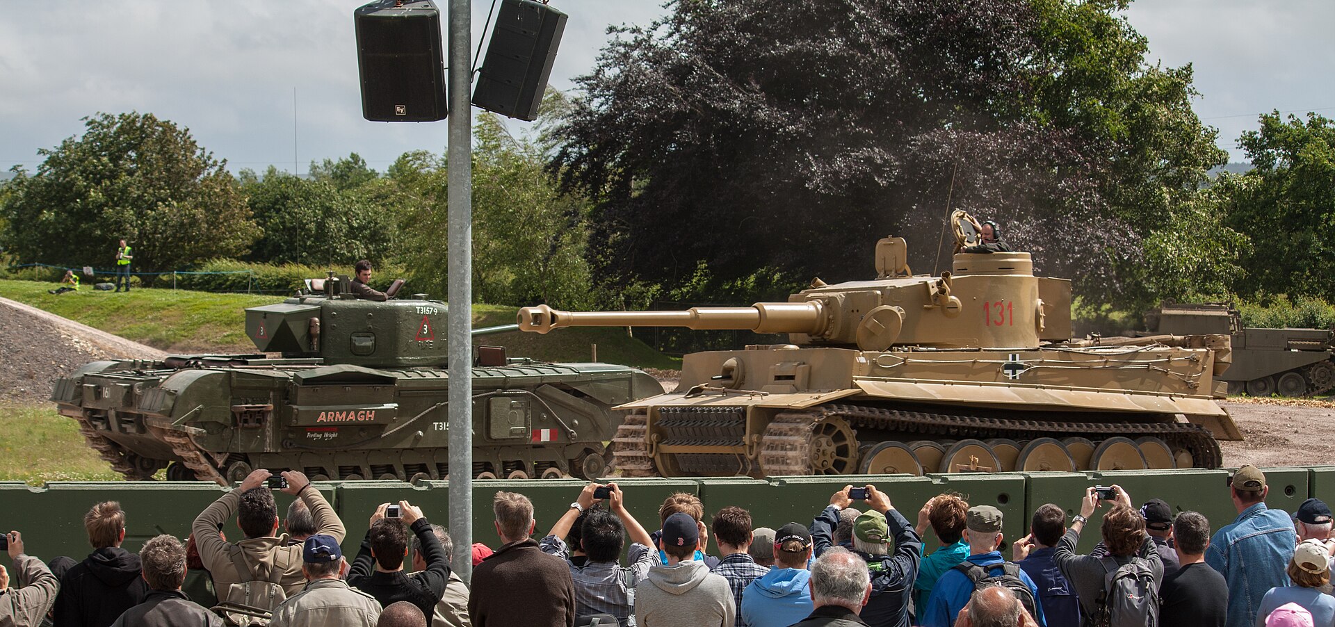 A Churchill tank and the Tiger 131 face each other in the Bovington arena while a crowd of spectators watches and takes photos