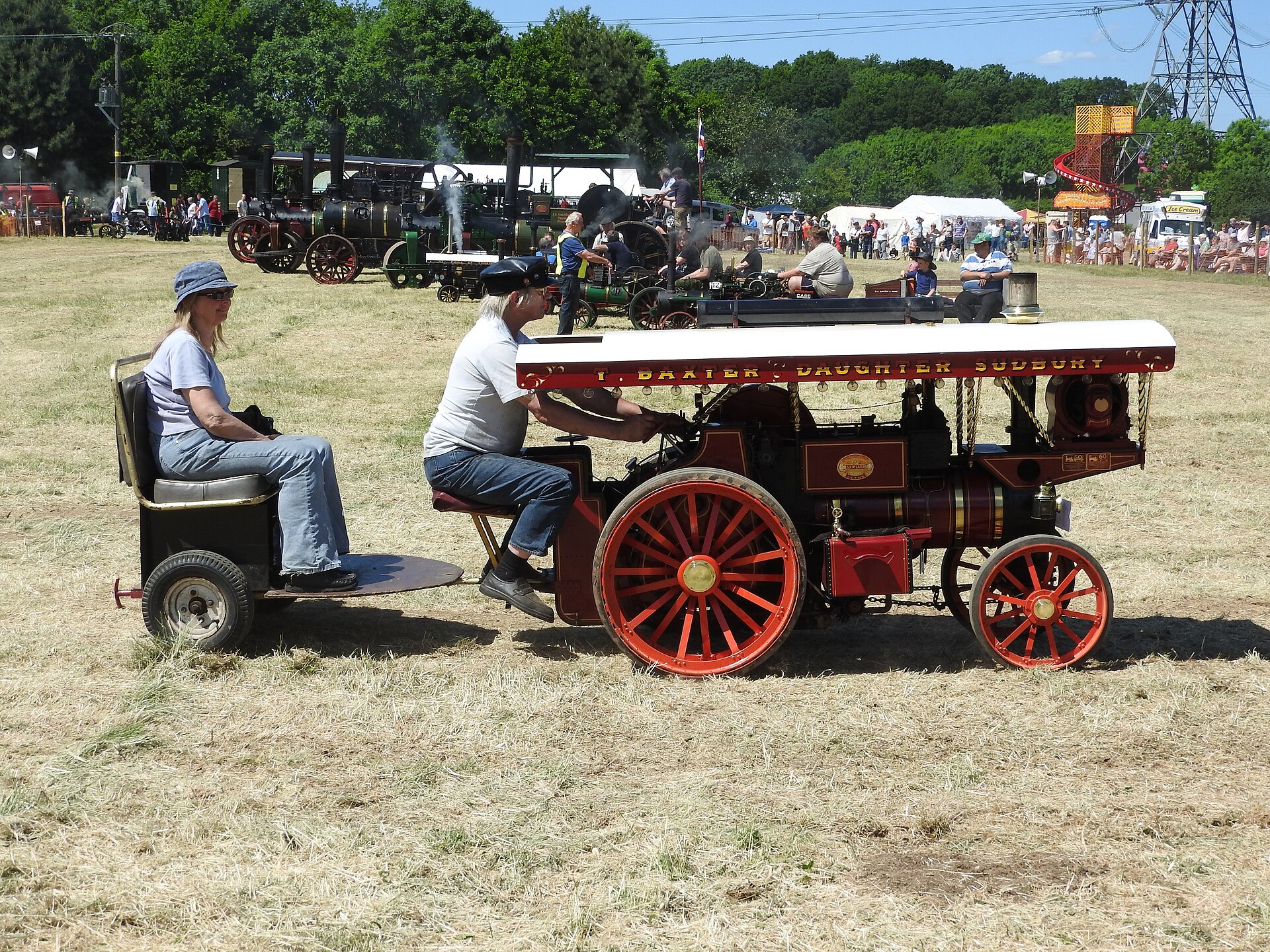 Couple riding a red-wheeled miniature showman's engine with white canopy across a sun-bleached rally field, full-size traction engines and a helter-skelter in the distance