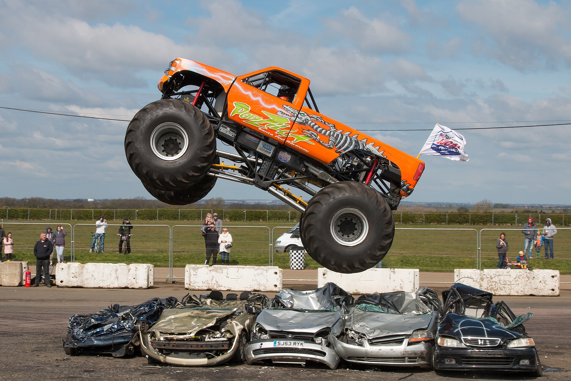 The orange Podzilla monster truck flies completely clear of a row of flattened cars, all four giant wheels off the ground under a blue spring sky.