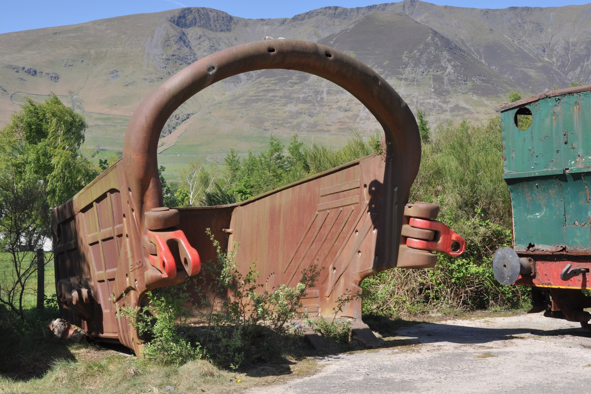A huge rust-brown dragline bucket taller than a person stands on a track at Threlkeld Quarry, its toothed lip and red shackles weathered, with the fells of Blencathra rising behind.
