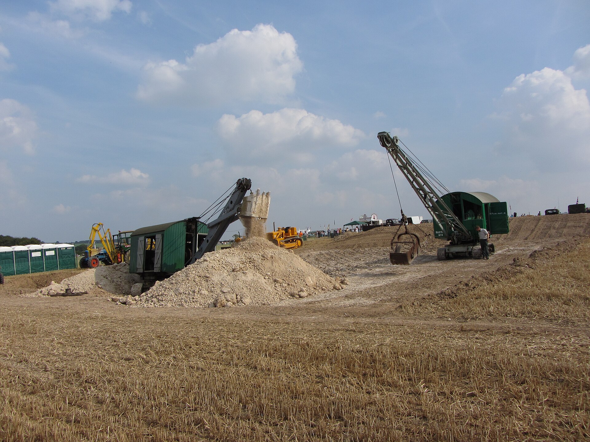 Two green Ruston-Bucyrus cable excavators work a mound of white chalk in a stubble field at the Great Dorset Steam Fair, one dropping its dragline bucket while an operator stands beside the other, beneath towering cumulus clouds.