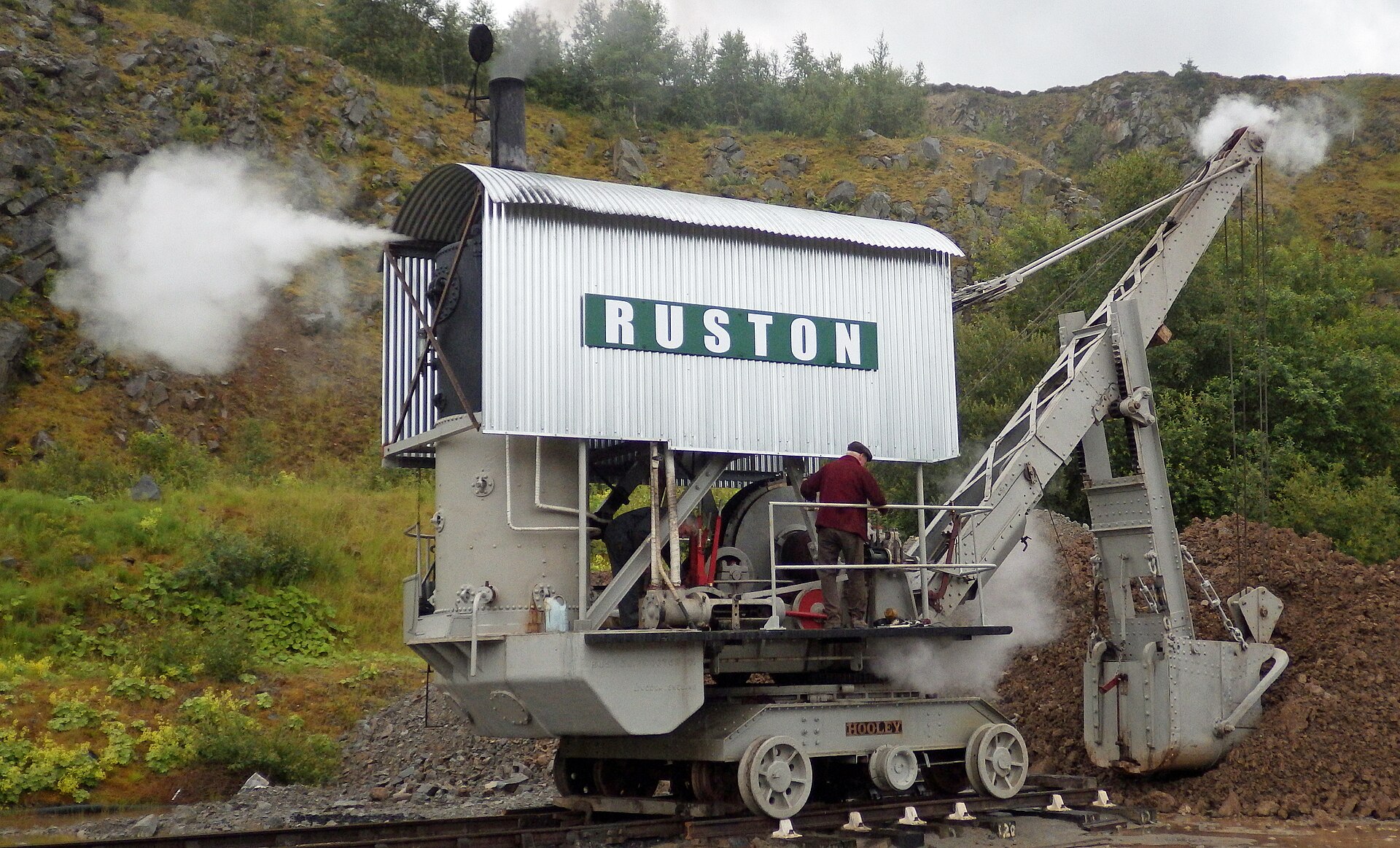 The silver corrugated 1909 Ruston steam excavator vents a horizontal jet of white steam while its operator in a red jumper works the controls, the riveted bucket resting against a bank of spoil.