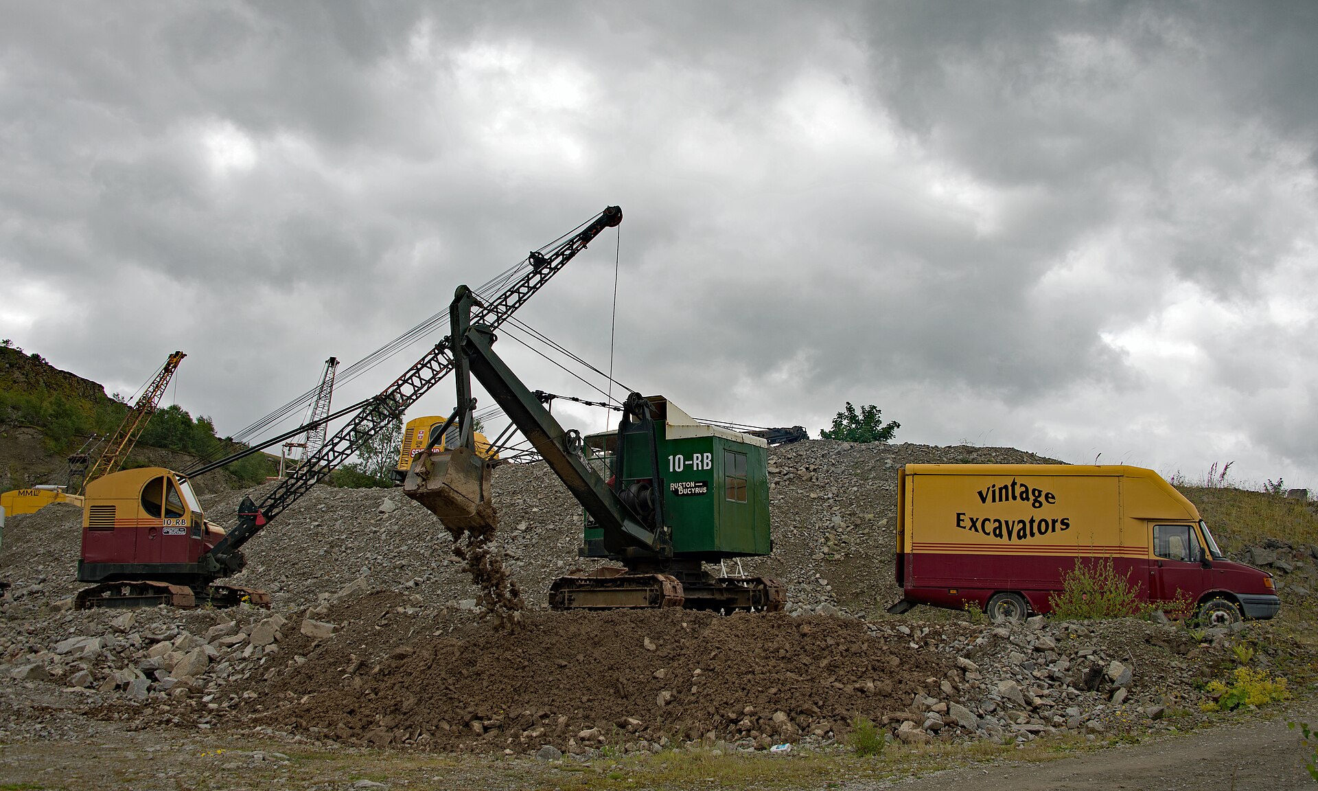 A green Ruston-Bucyrus 10-RB excavator tips spoil from its bucket in a rocky quarry under a brooding grey sky, with a vintage crane behind and a red-and-yellow Vintage Excavators van parked alongside.