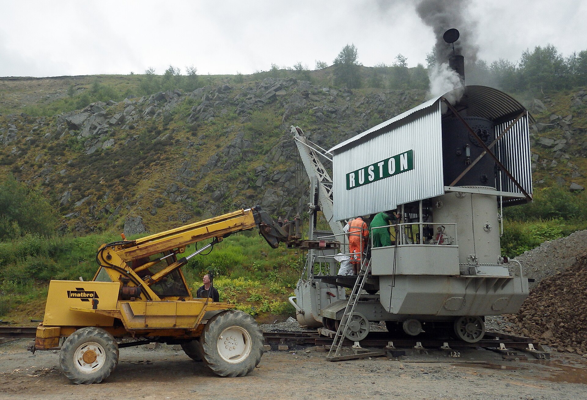 Crew in orange and white overalls load coal onto the corrugated-clad 1909 Ruston steam excavator as black smoke pours from its chimney, with a yellow Matbro loader alongside below a rocky hillside.
