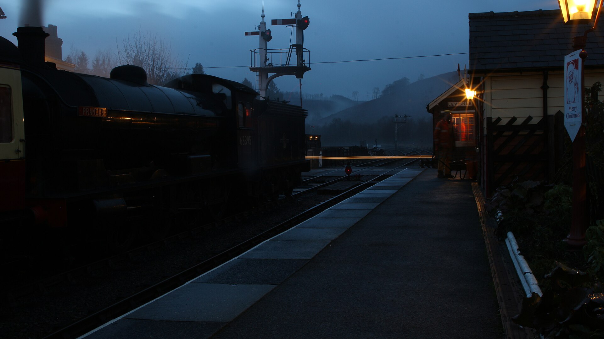 A black steam locomotive waits at a lower quadrant signal at Levisham station in low December light, steam drifting across the platform.