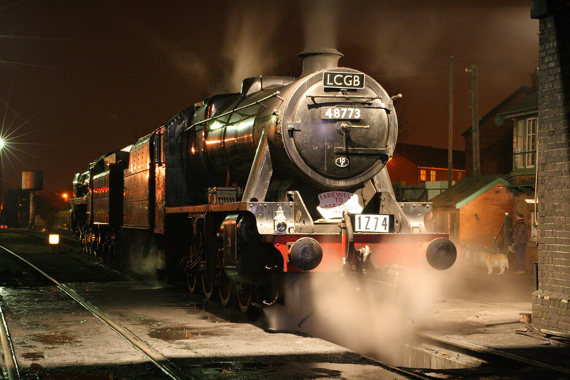 A blackened freight steam locomotive stands over the pit at Loughborough engine shed at night, lit by shed lamps, with another locomotive glowing in the dark behind.