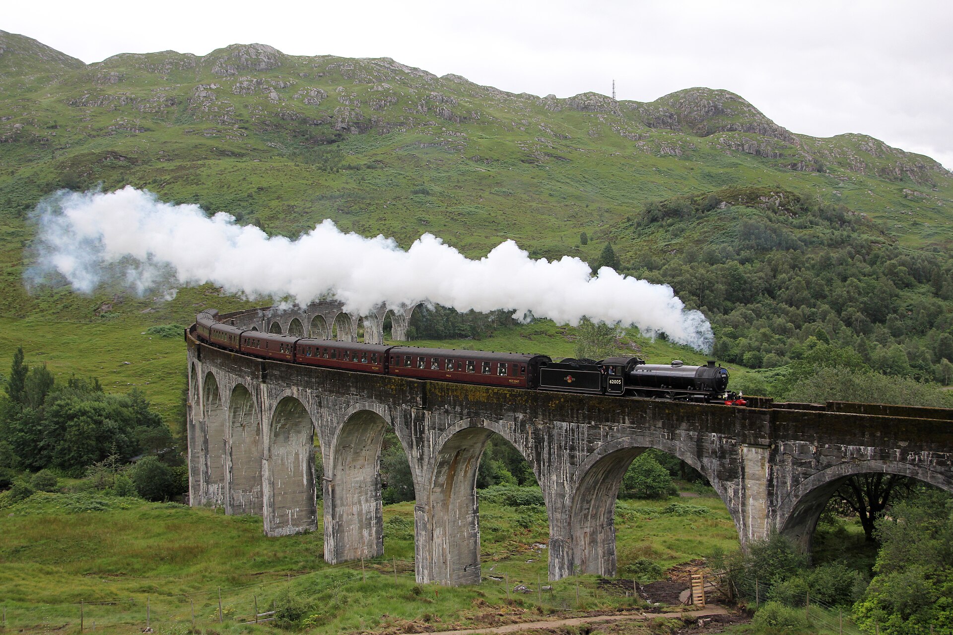 A steam locomotive hauls the Jacobite train across the curved stone arches of Glenfinnan Viaduct, white exhaust trailing back over the maroon carriages with Highland hills beyond.