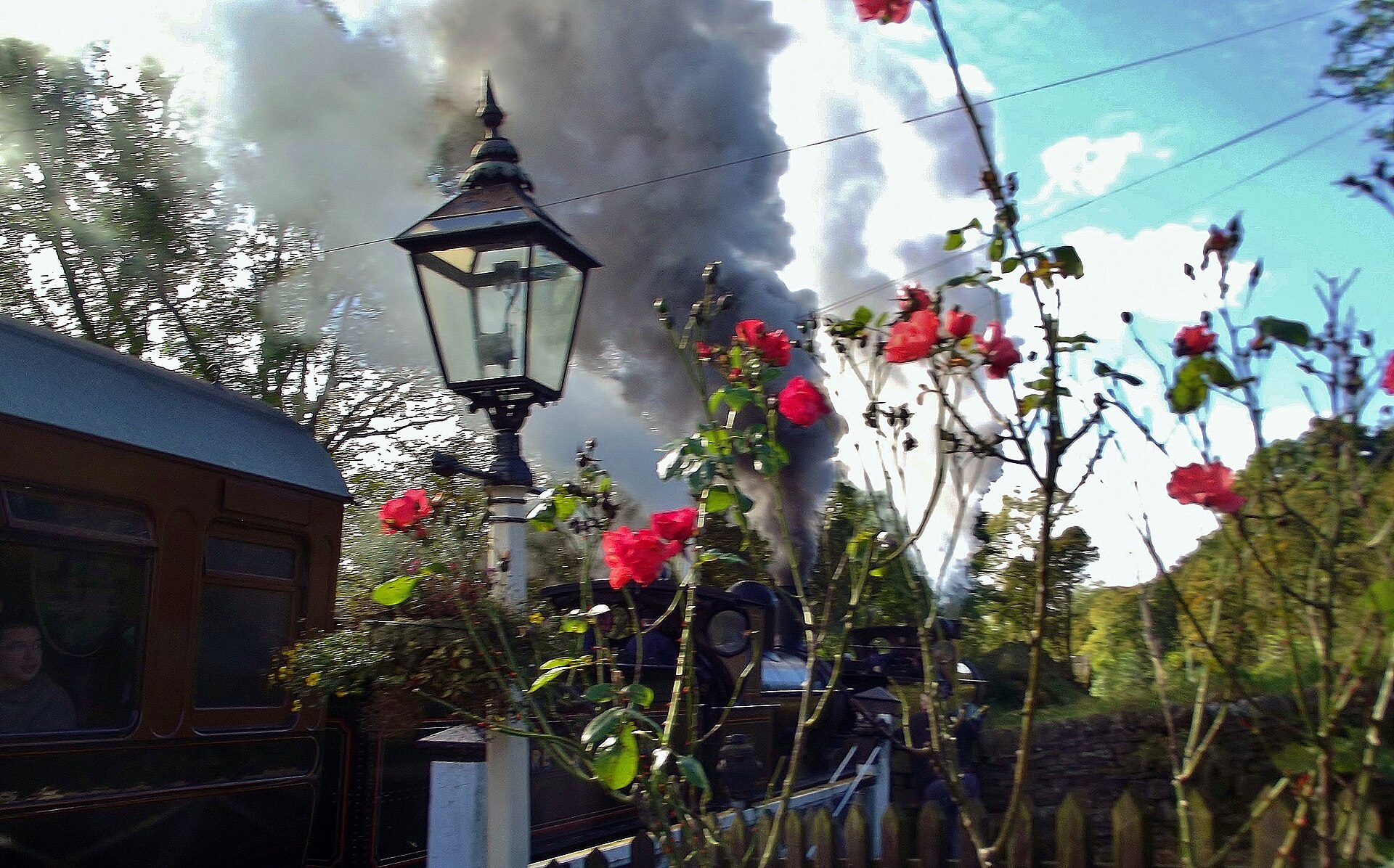 A steam locomotive pauses at Oakworth station on the Keighley and Worth Valley Railway, its gas-lit period platform framed by climbing roses.