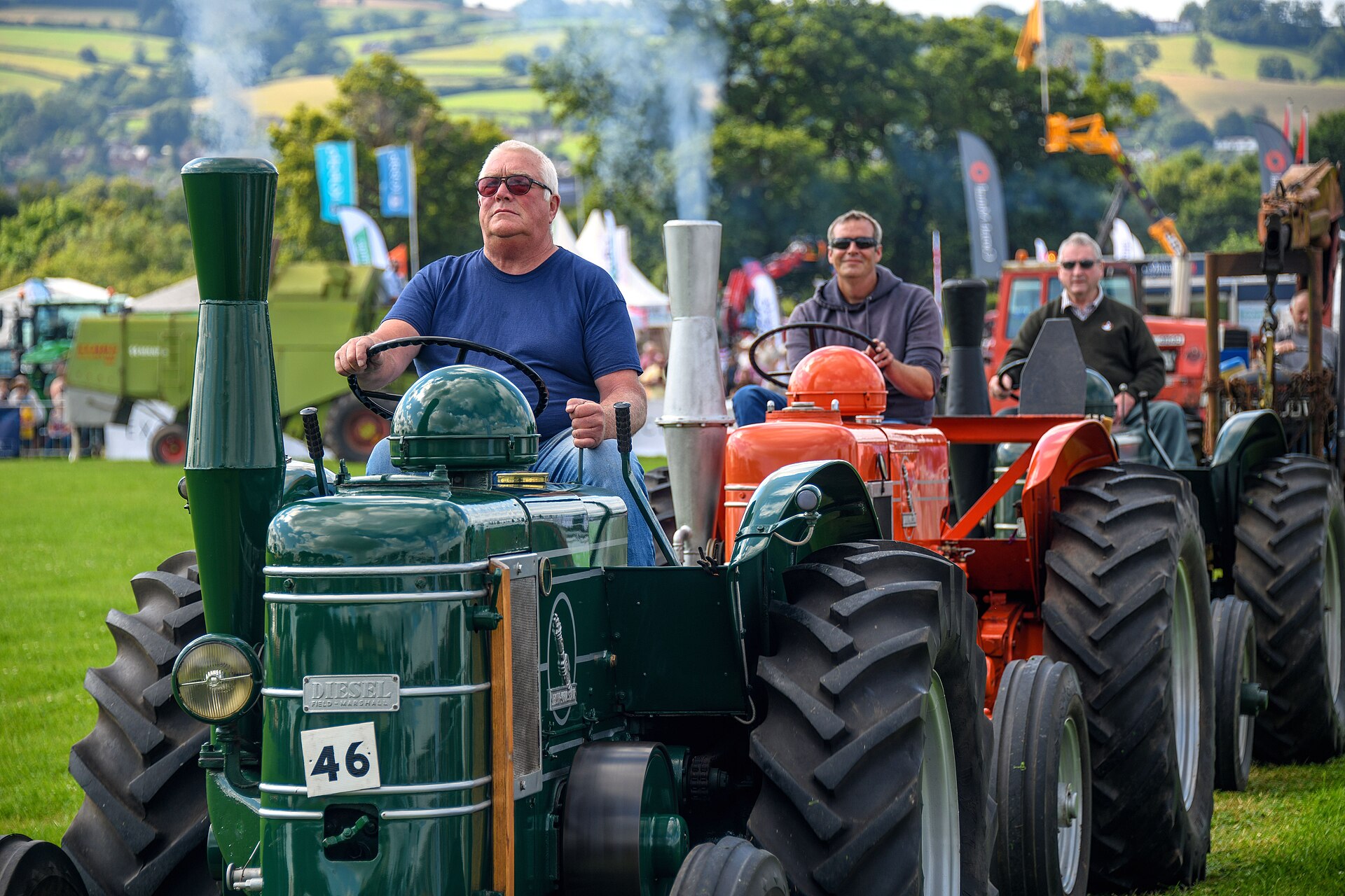 A driver on a green Field Marshall tractor numbered 46 leads a parade of vintage tractors at the Mid Devon Show, with exhaust smoke rising and Devon hills behind.