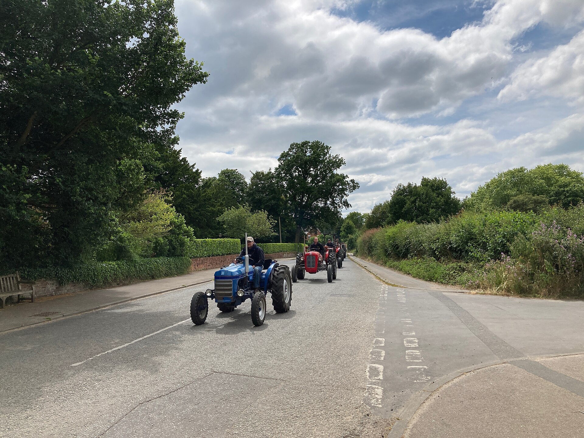 A convoy of vintage tractors led by a blue Fordson Dexta and a red Massey Ferguson drives through the village of Lavenham in Suffolk under a bright, cloud-filled sky.