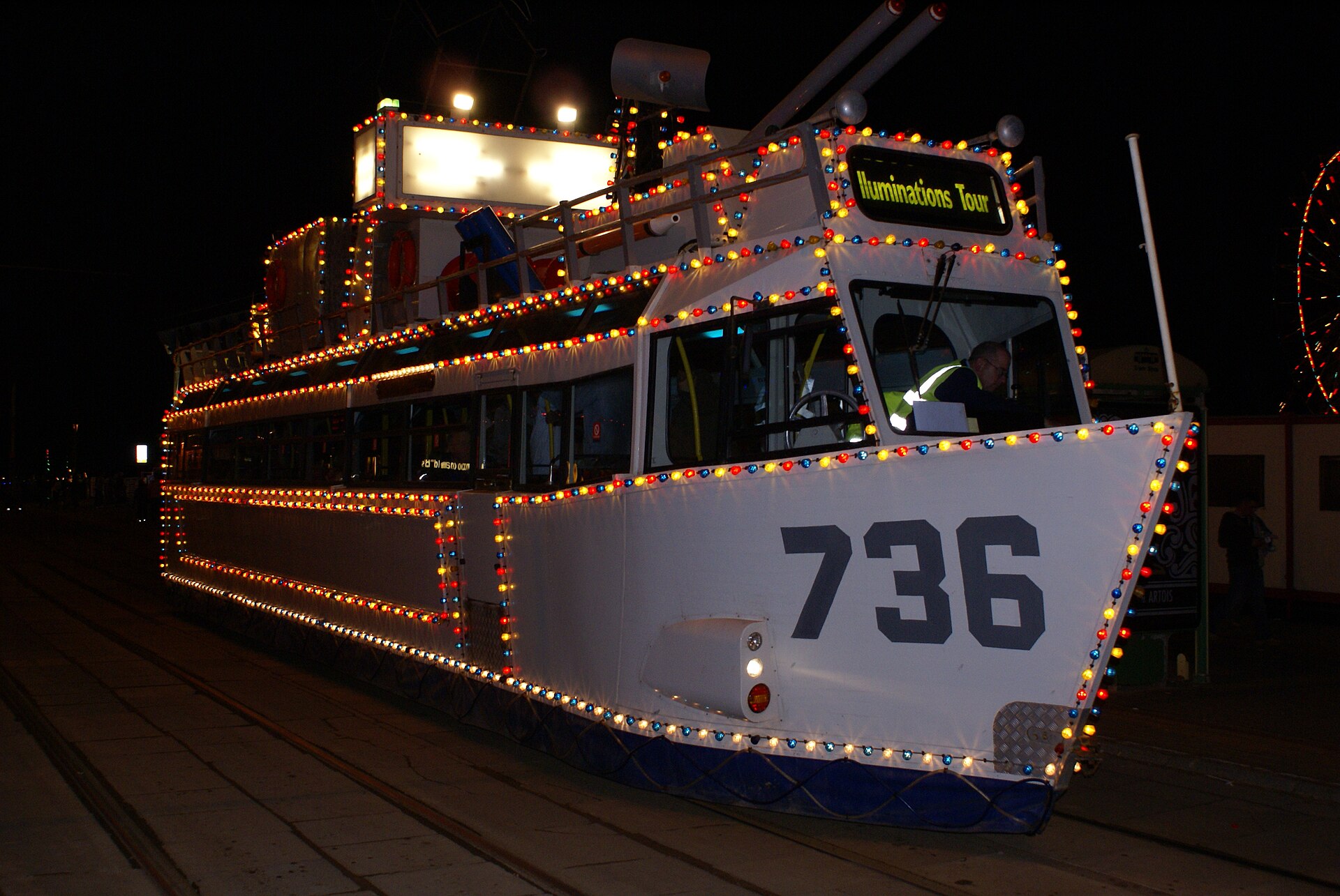 Blackpool's warship-shaped illuminated tram 736 covered in fairground bulbs at night, driver at the wheel under the Illuminations Tour sign