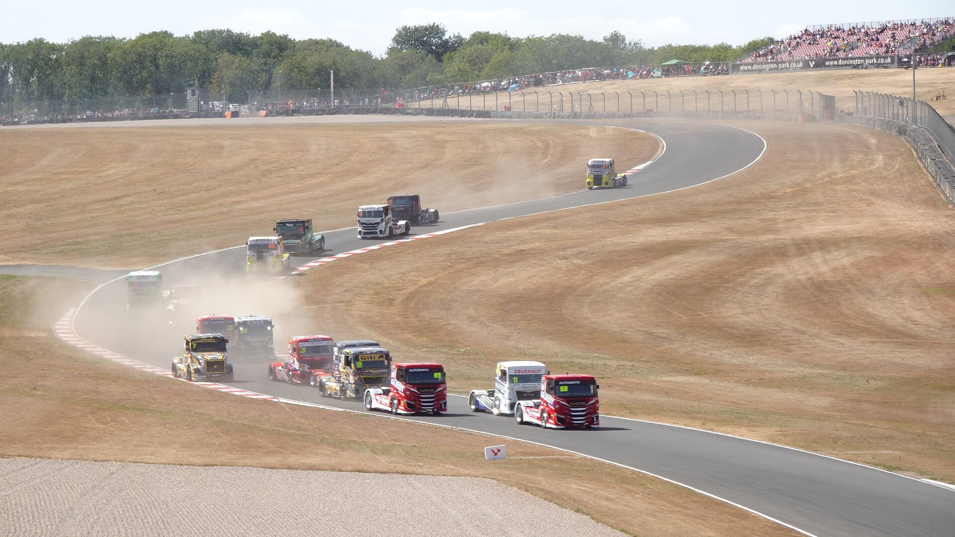 A field of racing trucks thunders downhill through Donington Park's Craner Curves towards the Old Hairpin on the first lap, kicking up dust with crowds lining the sun-parched banking.