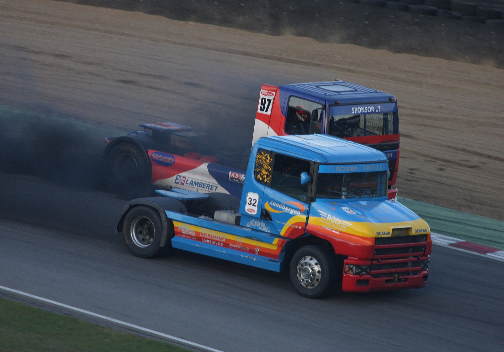 A blue, yellow and red Scania T-cab racing truck leads a rival through a corner at the Brands Hatch Truck Superprix, thick dark exhaust smoke trailing across the circuit.